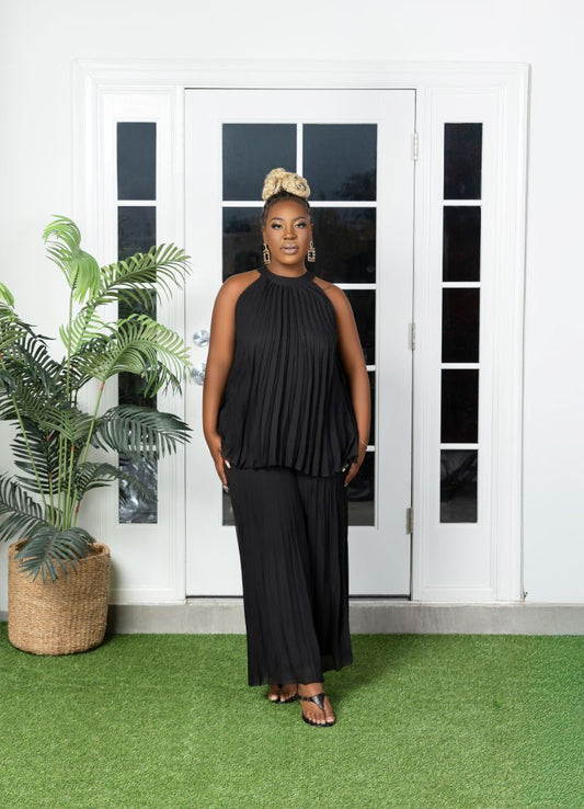 Woman in a black pleated outfit with a neck closure standing in front of a white door.