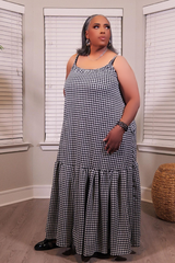 A woman modeling a sleeveless black and white houndstooth maxi dress with spaghetti straps and a tiered ruffle hem, standing in a room with window blinds.