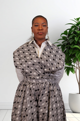 A woman wearing a gray wrap-style jumpsuit with a black and white circular pattern, layered over a white collared shirt, standing indoors next to a potted green plant.