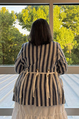 A woman viewed from behind wearing a blue and white vertically striped jacket with tie details at the waist, standing on a deck in front of a large window with green trees outside.
