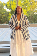 A woman stands outdoors wearing a navy and white vertical striped blazer over a white tiered maxi dress. The background shows a modern glass railing and green trees.