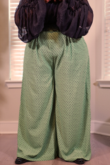 A person wearing wide-leg sage green pants with a small black polka dot pattern, paired with a sheer black puff-sleeve top, standing indoors in front of window blinds.
