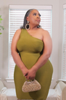 A woman wearing a one-shoulder olive green ribbed dress, holding a small beaded clutch purse in front of a window with white blinds.