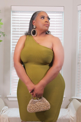 A woman wearing a one-shoulder olive green ribbed dress, holding a small beaded clutch purse in front of a window with white blinds.