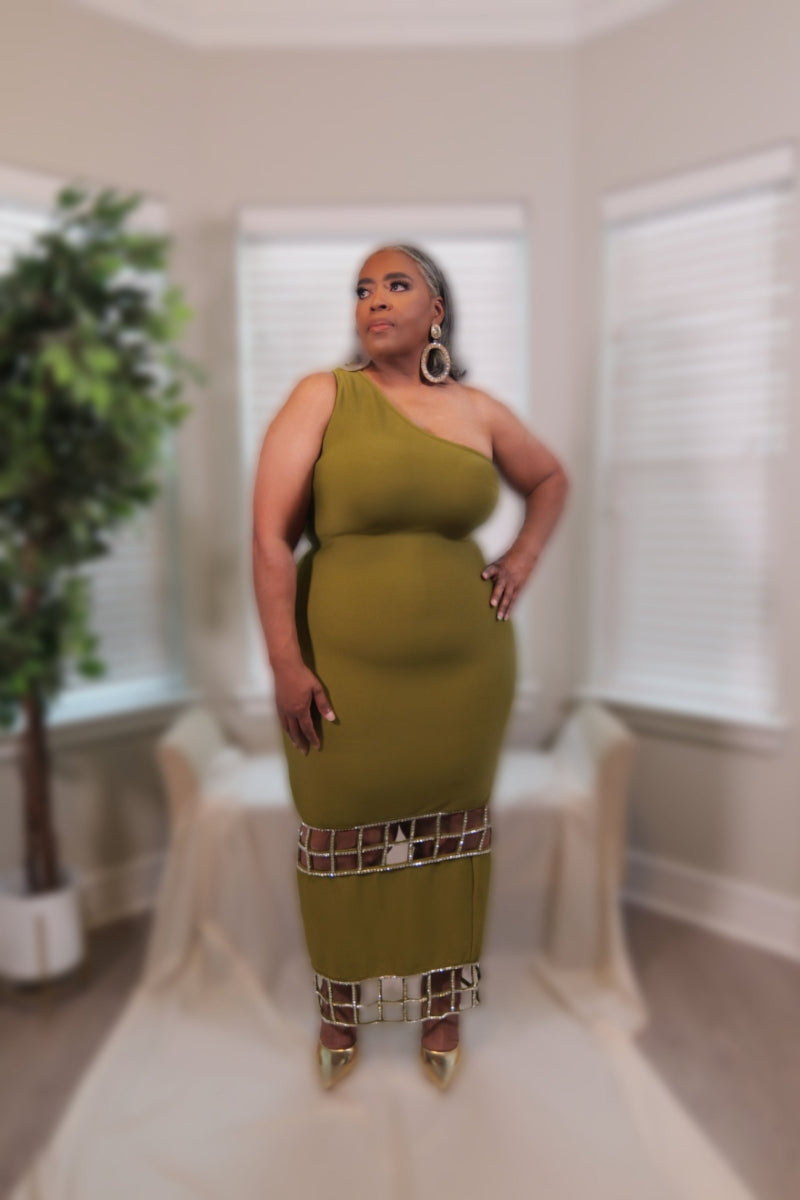 A woman modeling a one-shoulder olive green dress with a geometric cutout pattern near the hem, standing in a softly blurred indoor room with white window blinds and a potted plant.
