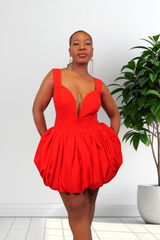 A woman modeling a bright red sleeveless mini dress with a deep plunging neckline and a voluminous gathered skirt, standing in a room with a white wall and a potted green plant.