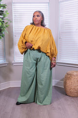 A woman modeling a two-piece outfit consisting of a mustard yellow off-the-shoulder long-sleeve top and sage green wide-leg pants with a small white polka dot pattern, standing in a room with window blinds.