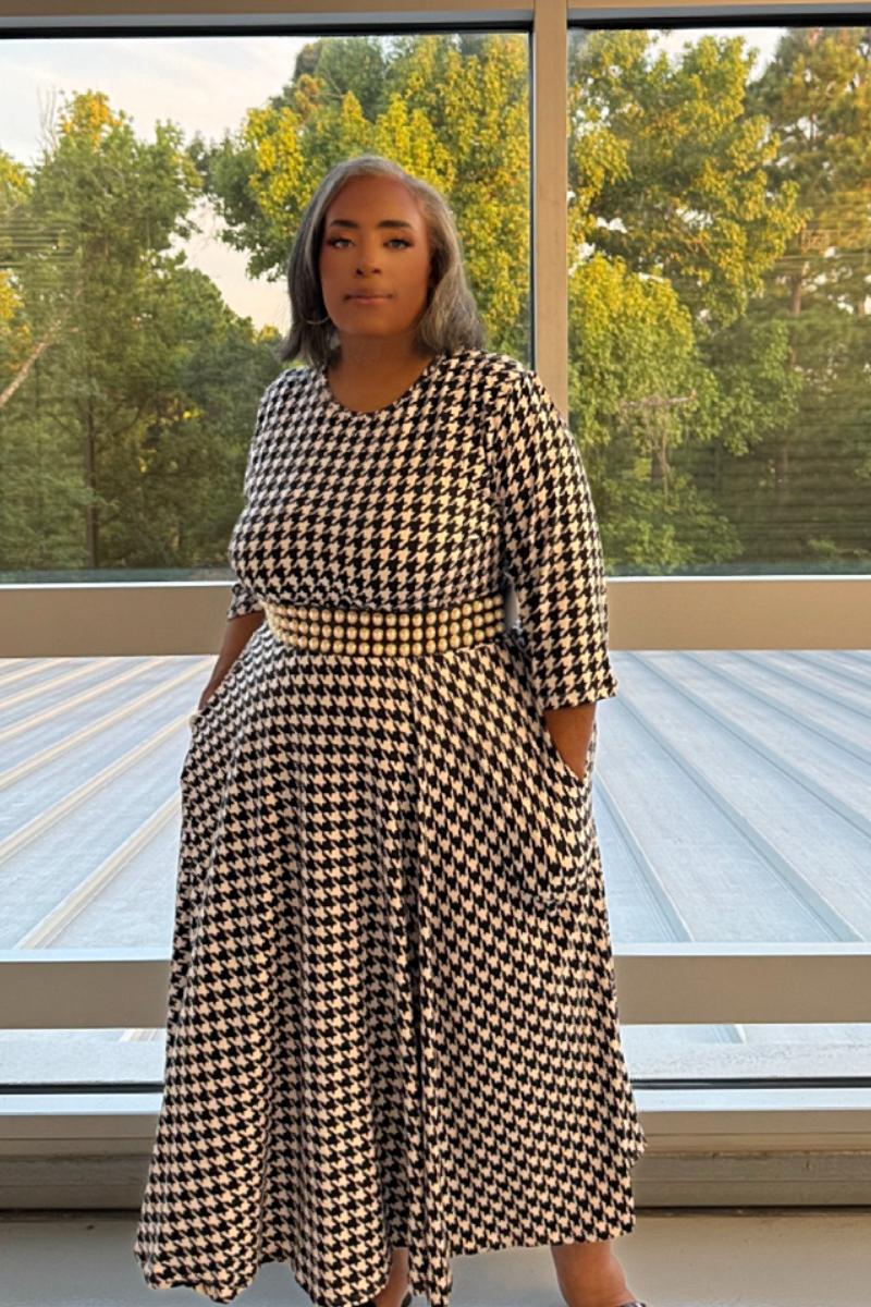 A woman modeling a black and white houndstooth midi dress with elbow-length sleeves and a matching waist belt, standing in front of a large window with trees outside.