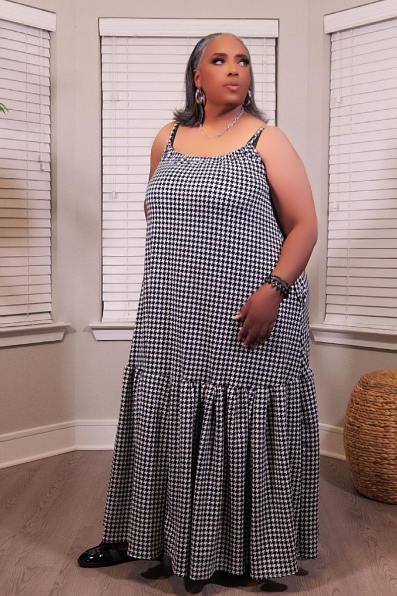 A woman modeling a sleeveless black and white houndstooth maxi dress with spaghetti straps and a tiered ruffle hem, standing in a room with window blinds.