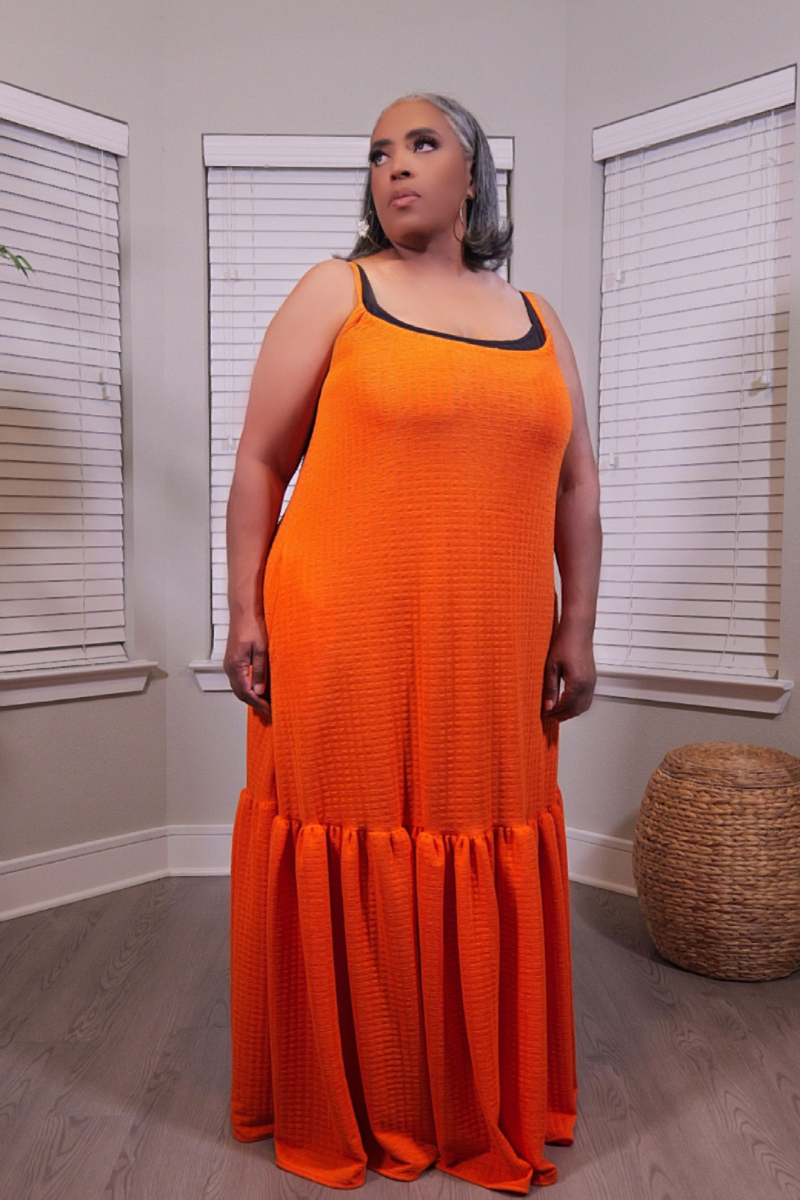A woman modeling a bright orange textured maxi dress with black trim on the straps and neckline, standing in a room with window blinds and a woven basket on the floor.