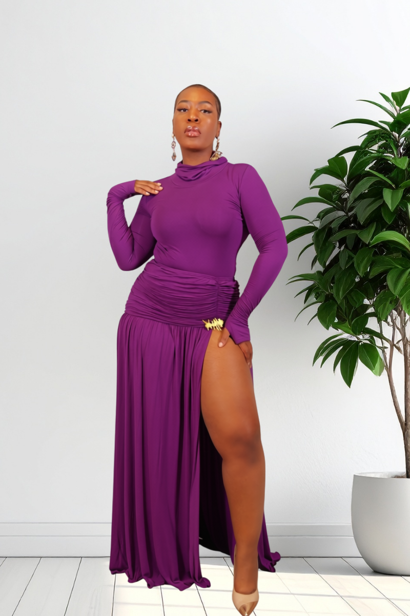 A woman modeling a long-sleeve, high-neck, floor-length purple dress with a draped waist and a thigh-high slit, standing in a bright room with a potted plant on the right.