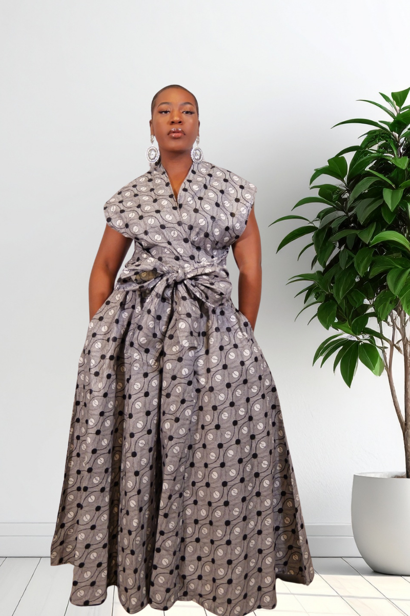 A woman modeling a sleeveless wrap-style maxi jumpsuit with a tied waist and a gray, black, and white circular pattern, standing in a bright room next to a potted plant.