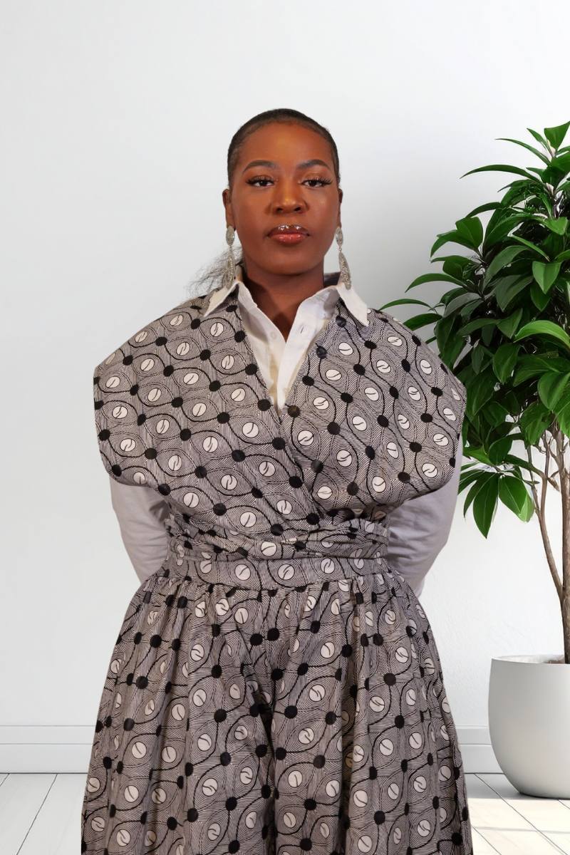 A woman wearing a gray wrap-style jumpsuit with a black and white circular pattern, layered over a white collared shirt, standing indoors next to a potted green plant.