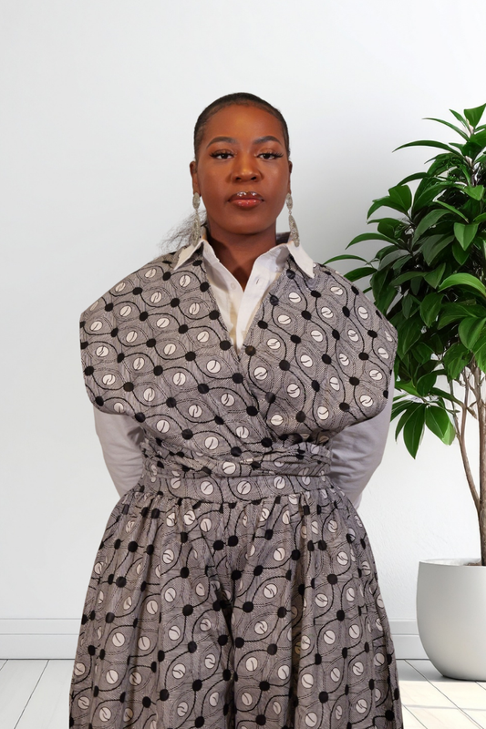 A woman wearing a gray wrap-style jumpsuit with a black and white circular pattern, layered over a white collared shirt, standing indoors next to a potted green plant.