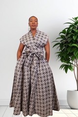 A woman modeling a sleeveless wrap-style maxi jumpsuit with a tied waist and a gray, black, and white circular pattern, standing in a bright room next to a potted plant.