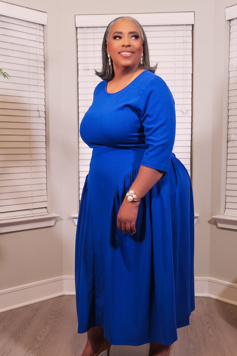 A woman modeling a bright royal blue midi dress with three-quarter sleeves and a gathered skirt, standing in front of white window blinds.