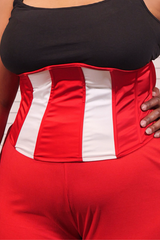 A close-up of a person wearing a red waist trainer or waist cincher with white vertical stripes, paired with a black top and red bottoms.
