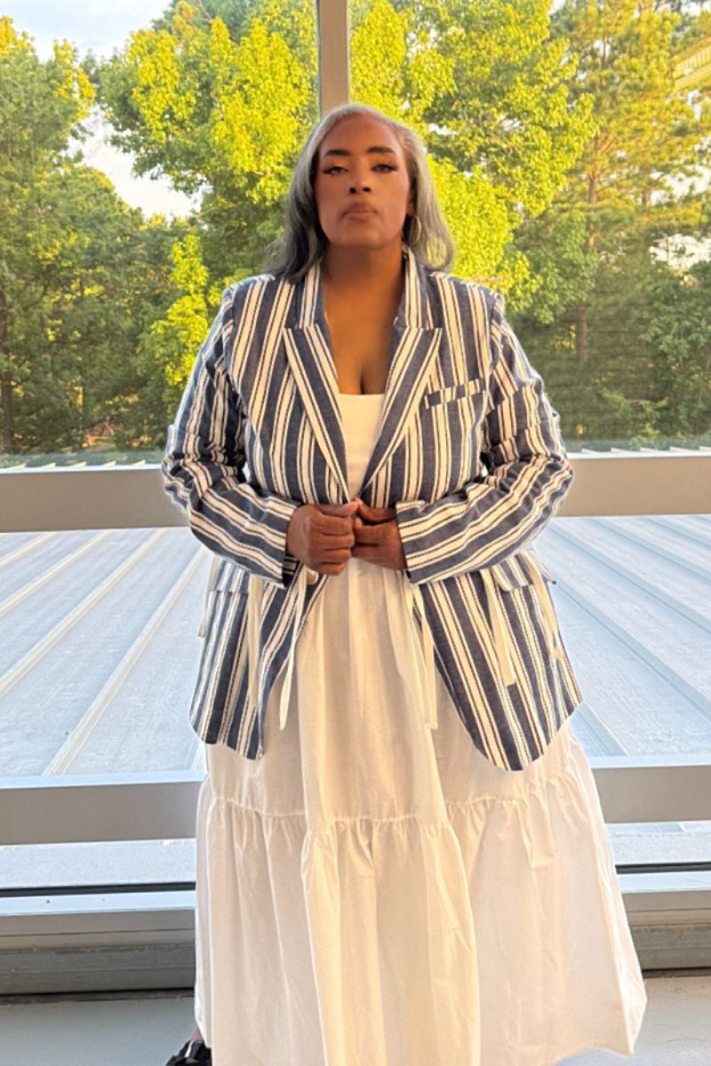 A woman stands outdoors wearing a navy and white vertical striped blazer over a white tiered maxi dress. The background shows a modern glass railing and green trees.