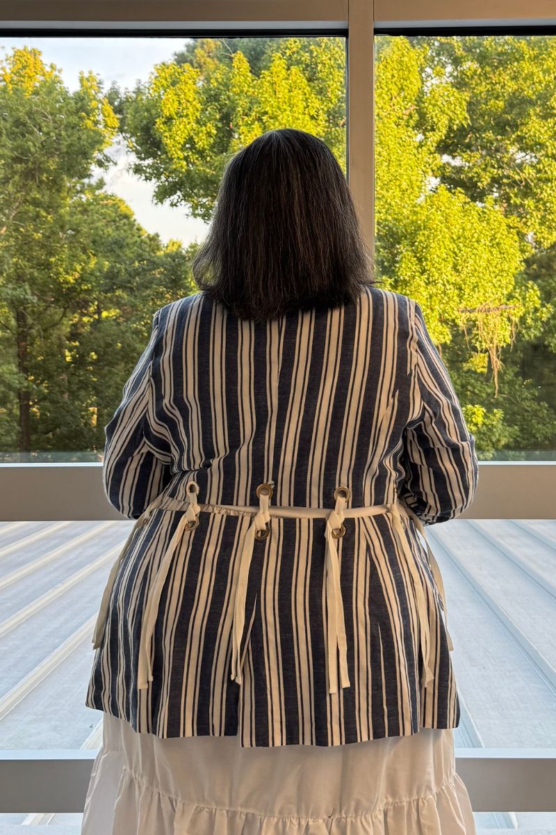 A woman viewed from behind wearing a blue and white vertically striped jacket with tie details at the waist, standing on a deck in front of a large window with green trees outside.