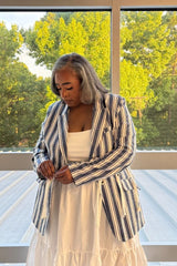 A woman with gray hair is wearing a navy and white vertically striped blazer over a white tiered dress, standing in front of a large window with green trees outside.