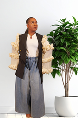 A woman modeling a black blazer with dramatic cream ruffled puff sleeves, paired with a white top and wide-leg blue jeans, standing next to a potted green plant against a white wall.