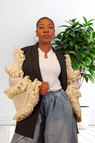 A woman wearing a black jacket with dramatic cream ruffled sleeves, a white top, and a blue jeans, standing in front of a white wall with a green potted plant.