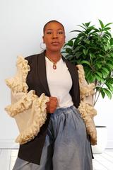 A woman wearing a black jacket with dramatic cream ruffled sleeves, a white top, and a blue jeans, standing in front of a white wall with a green potted plant.
