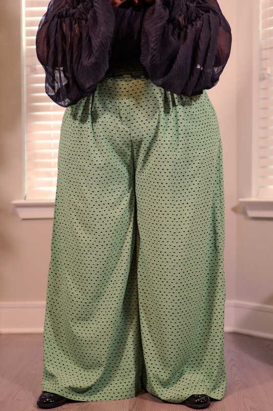 A person wearing wide-leg sage green pants with a small black polka dot pattern, paired with a sheer black puff-sleeve top, standing indoors in front of window blinds.
